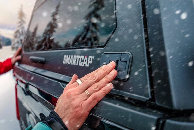 A hand presses the latch on a steel SmartCap truck canopy in falling snow.